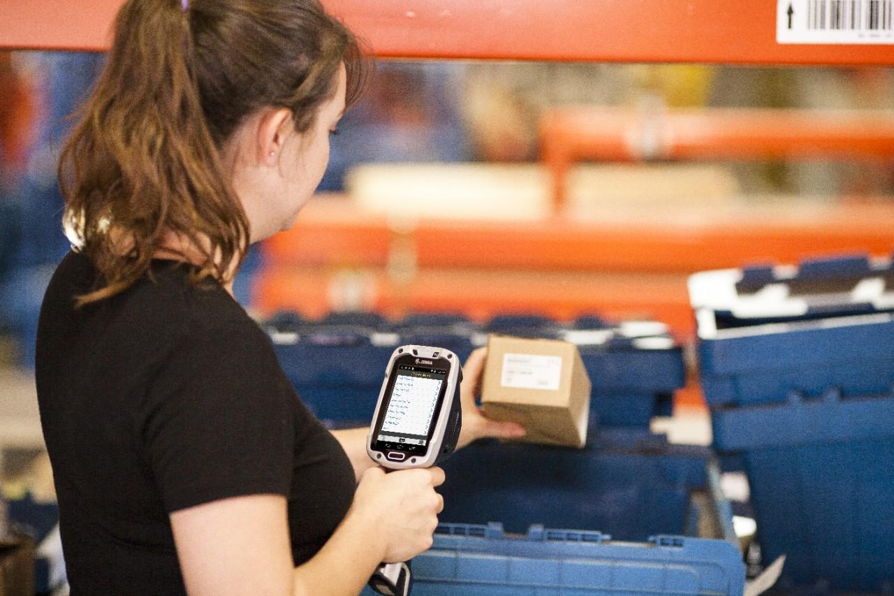 A warehouse worker holding a Zebra TC8000 in one hand and a small carton
                  in the other, in front of blue stock totes on orange racking